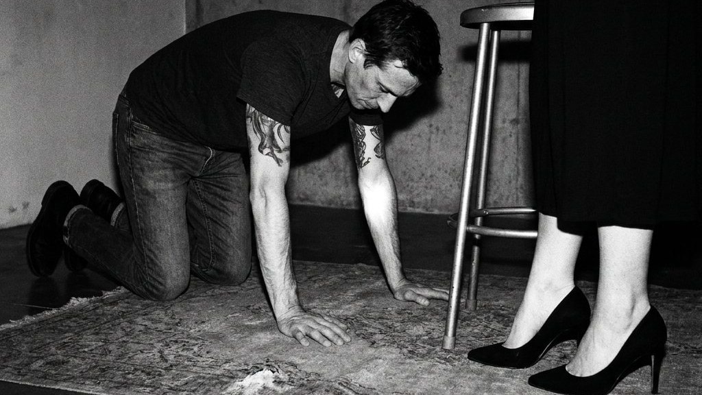 A man on his hands and knees on a rug beside a woman standing in black heels