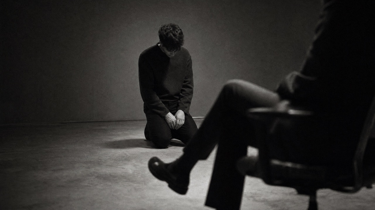 A person kneeling on the floor in a dim room, head bowed, beside the legs of someone seated and fully dressed