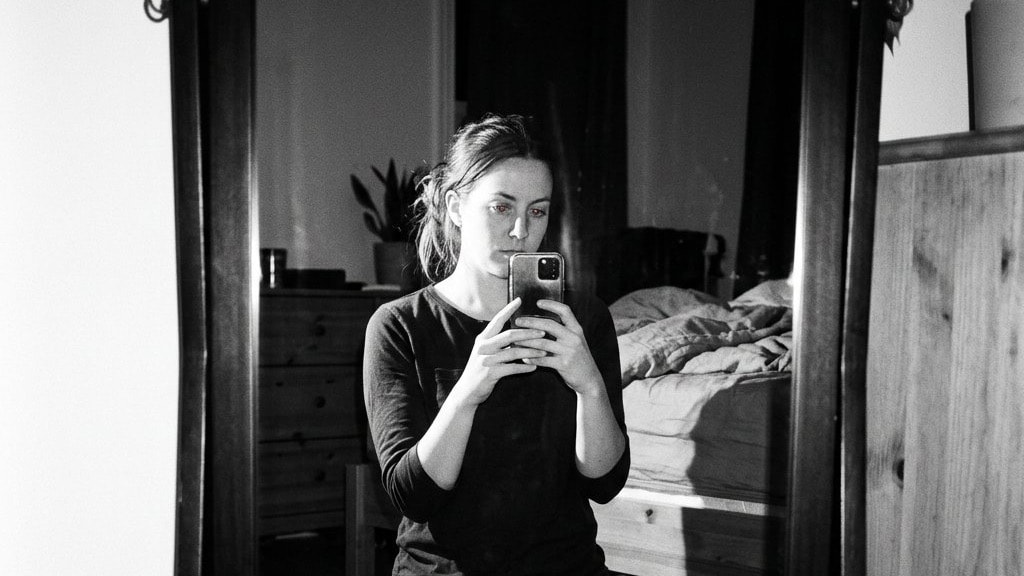 A woman kneeling in a bedroom, taking a mirror photo on her phone as proof